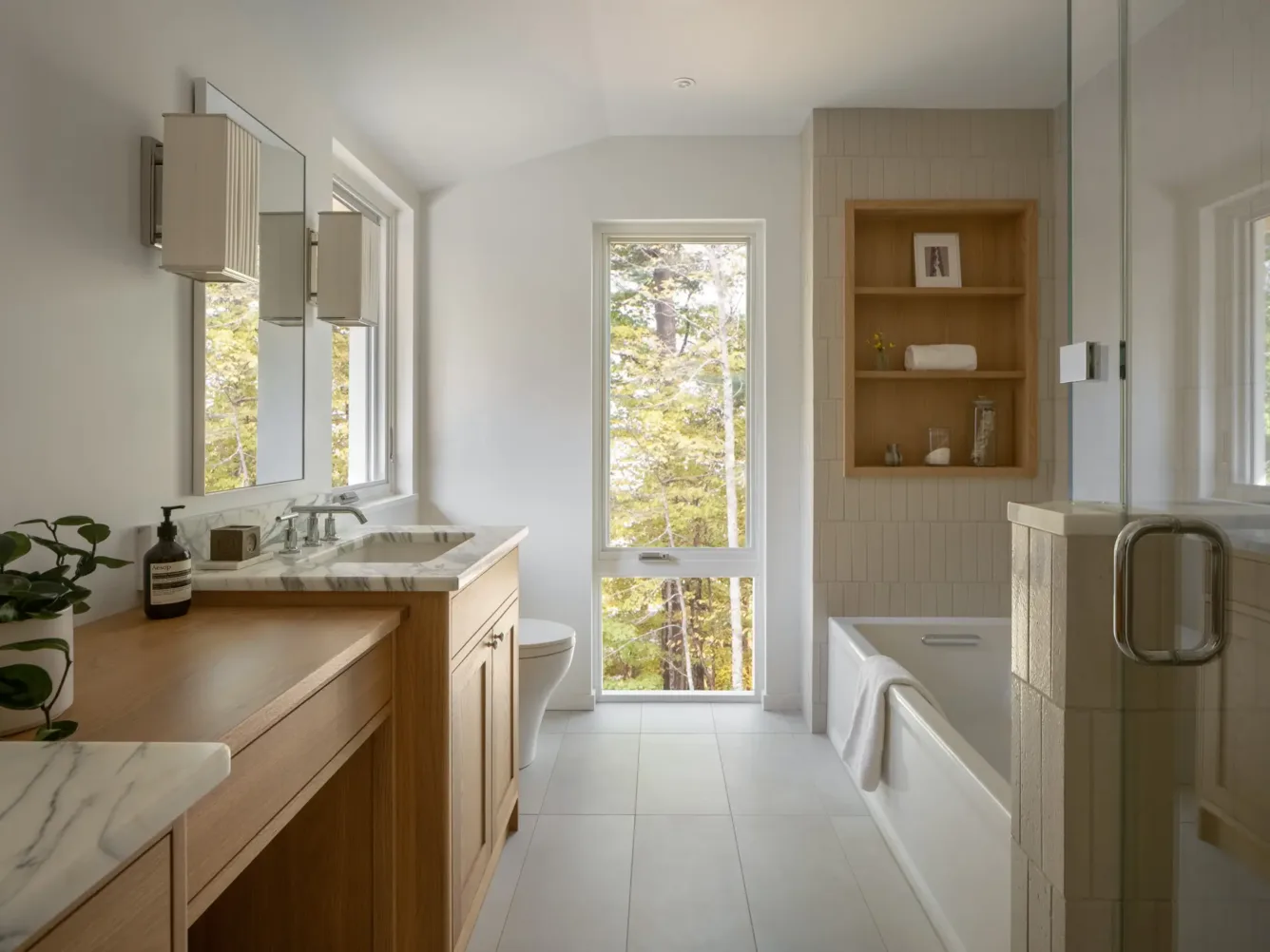 Modern bathroom with wood and marble accents, large window with trees outside, bathtub, glass shower, and neatly arranged towels and plants.