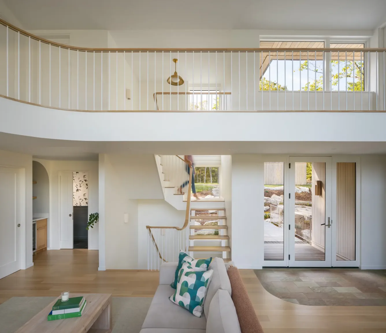 Bright, modern living room with open staircase, beige sofa, wooden floors, large windows, and a balcony railing overlooking the space. Glass doors lead outside.
