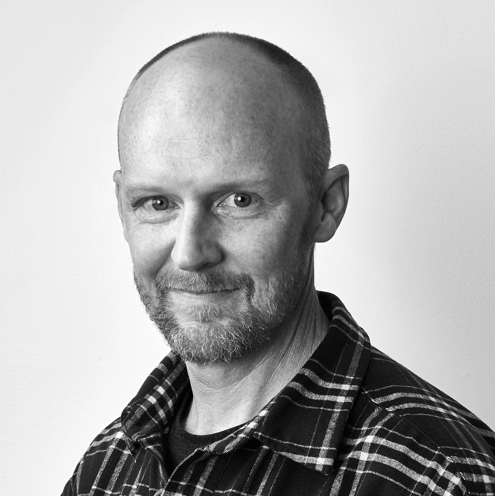 Black and white portrait of a middle-aged man with a beard and bald head, wearing a plaid shirt, smiling slightly, and looking at the camera against a plain background.
