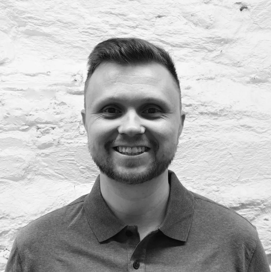 A man with short hair and a beard smiles at the camera, wearing a collared shirt, standing in front of a textured white brick wall.