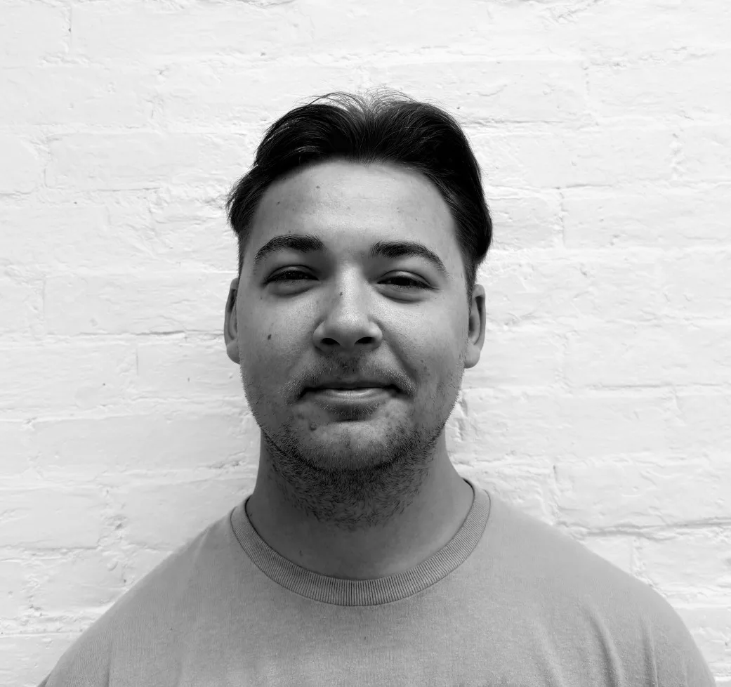 Young man with short dark hair and slight facial hair, smiling softly, stands in front of a white brick wall. Black and white photo.