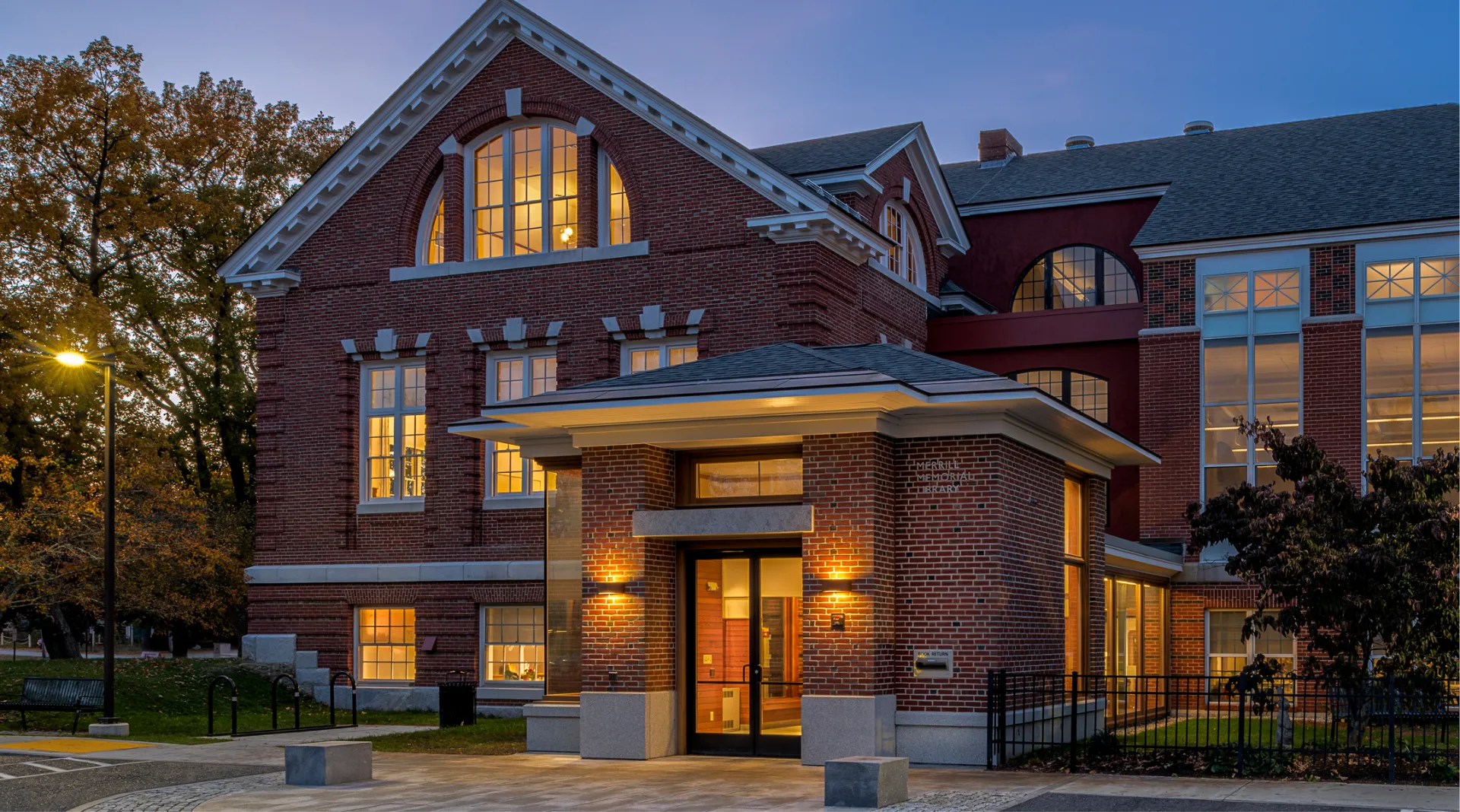 A red-brick building with large windows warmly lit from within, photographed at dusk with trees and a lamp post visible outside.
