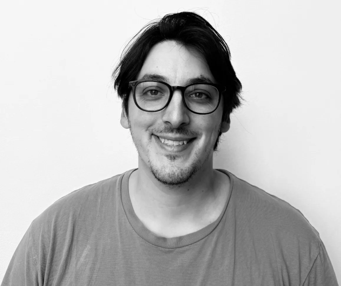 A man with dark hair, glasses, and a short beard smiles at the camera. He is wearing a T-shirt and standing against a plain, light-colored background.