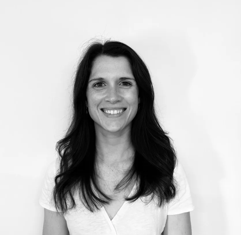 A woman with long dark hair and a white shirt smiles at the camera in front of a plain white background.