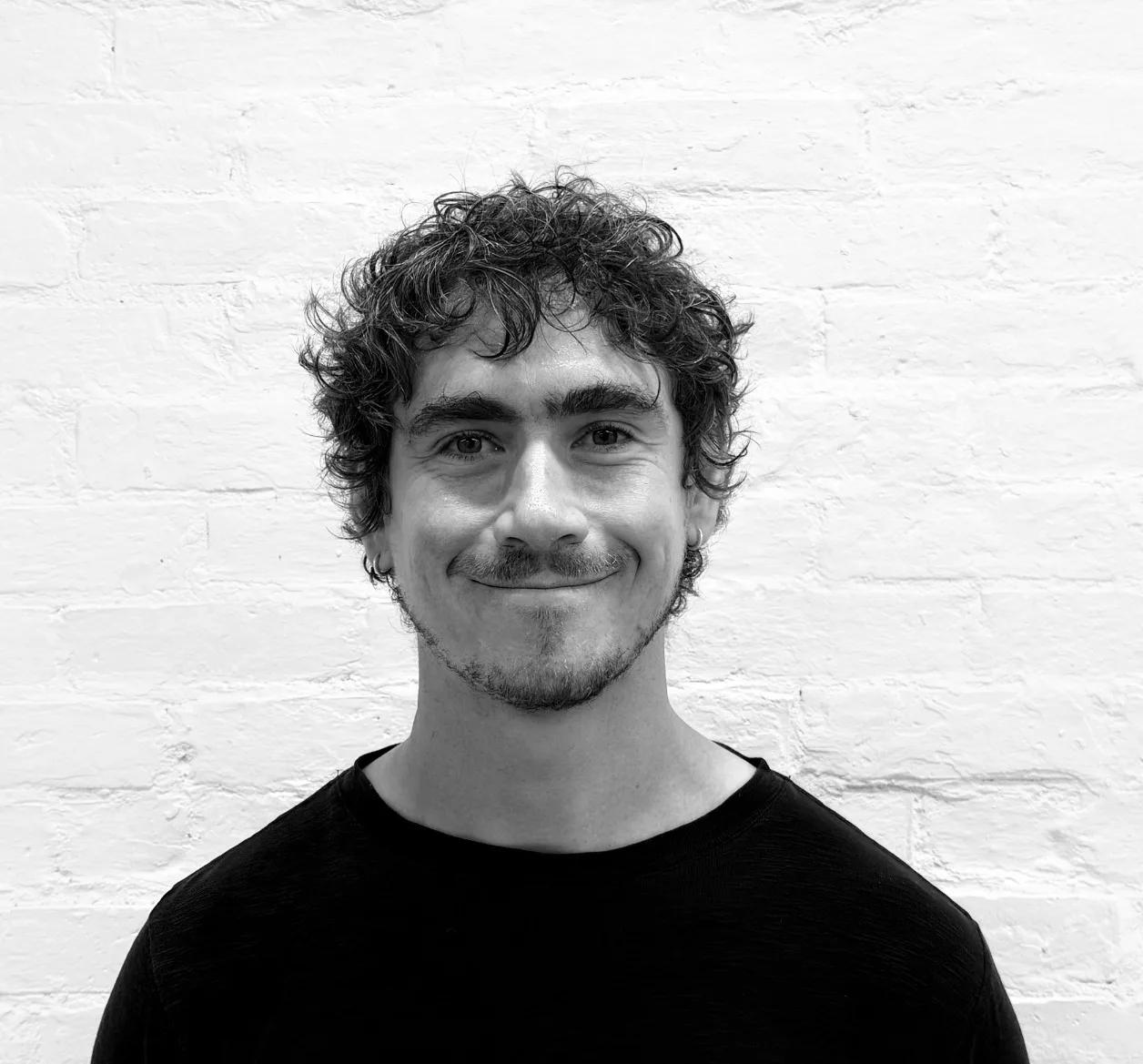 Man with curly hair and a short beard smiles in front of a white brick wall, wearing a dark t-shirt. The photo is in black and white.