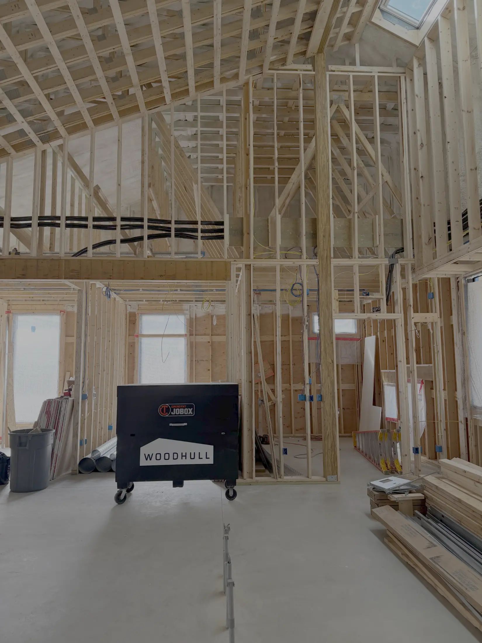 Interior of a house under construction, showing exposed wooden framing, a skylight, building materials, and a black tool chest labeled “WOODHULL” on a concrete floor.