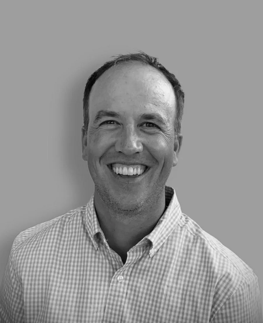 Smiling man in a checkered button-up shirt against a plain gray background, shown from the shoulders up, in black and white.