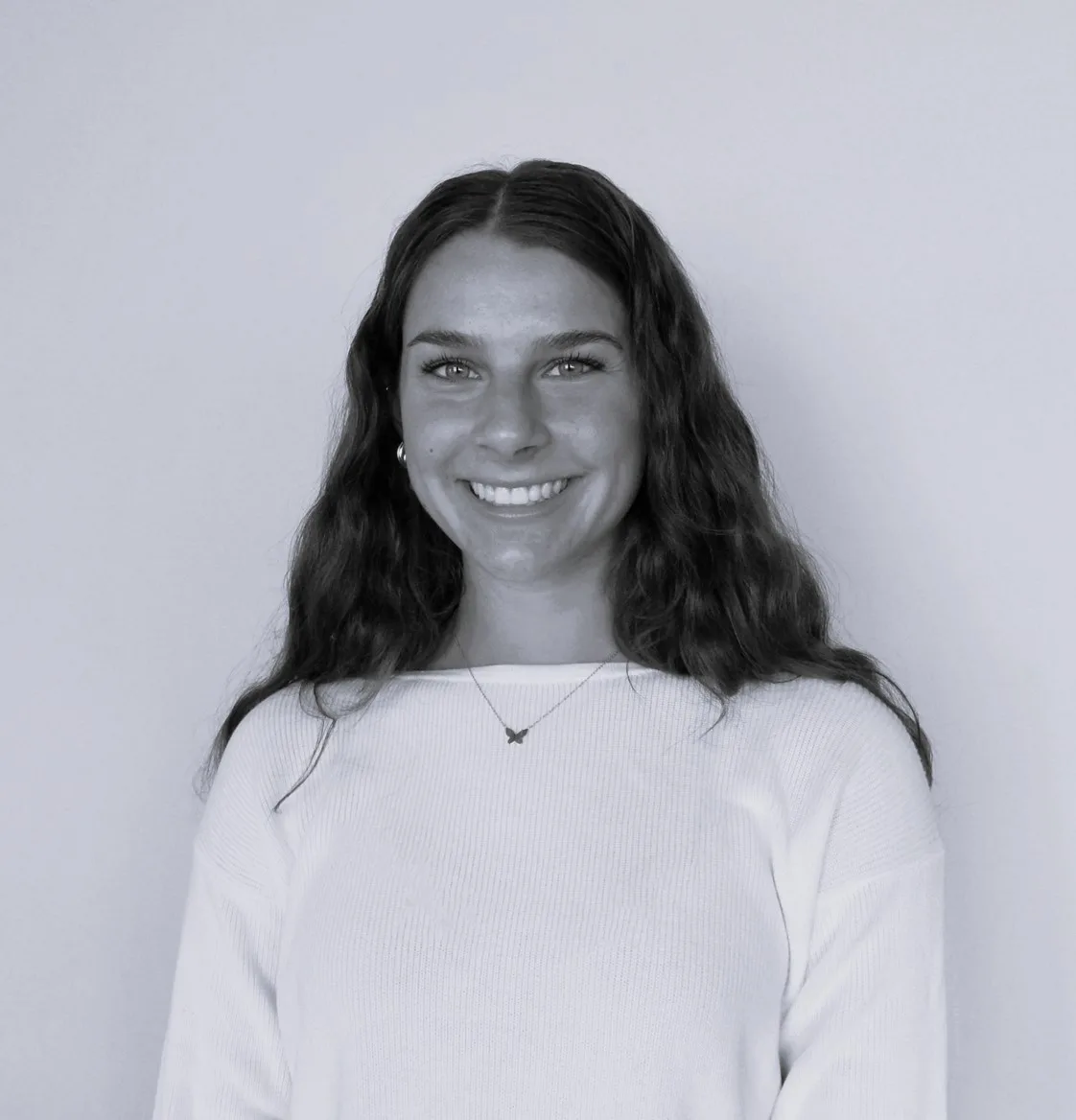 A young woman with long wavy hair smiles at the camera. She is wearing a light-colored long-sleeve top and a necklace, standing against a plain background.