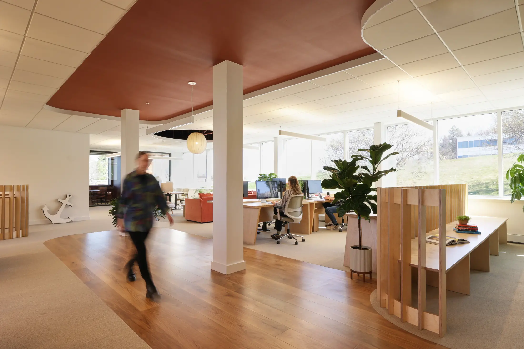 Modern open-plan office with desks, computers, plants, and large windows; one person walks while others work at their desks in a bright, well-lit space.