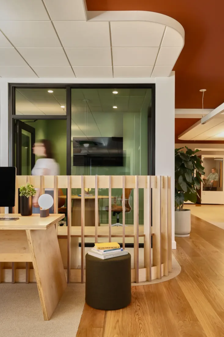 Modern office space with wood furniture, potted plants, a computer on a desk, glass-walled meeting room, and people walking in blurred motion. Warm lighting and earthy tones throughout.