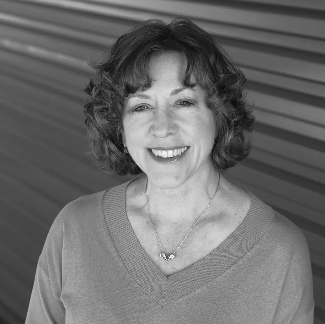 Smiling woman with curly hair wearing a sweater and necklace, standing in front of a wall with horizontal lines. Black and white photo.