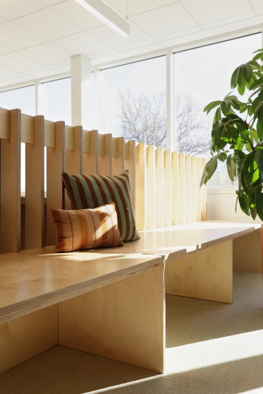 A wooden bench with striped cushions is positioned by large windows, allowing sunlight to stream in. A green leafy plant sits nearby, creating a bright and cozy atmosphere.