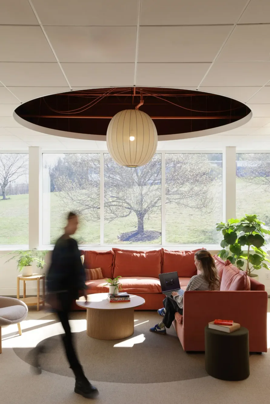 A person walks past another person sitting on a coral sectional sofa with a laptop in a bright lounge with large windows, a round hanging light, and indoor plants.