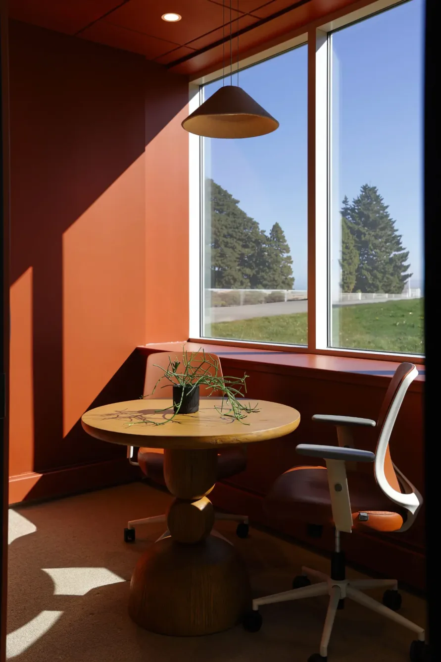 A small office with red walls, a round wooden table, two chairs, a hanging lamp, and a potted plant by a large window letting in sunlight. Trees are visible outside.