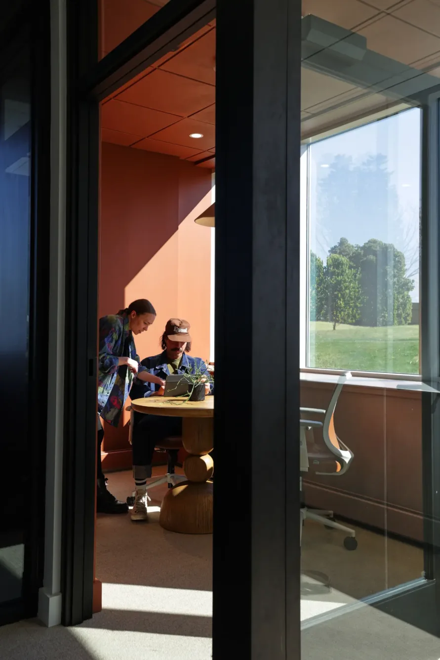 Two people work together at a small round table in a sunlit office with orange walls and large windows, while an empty chair sits nearby.