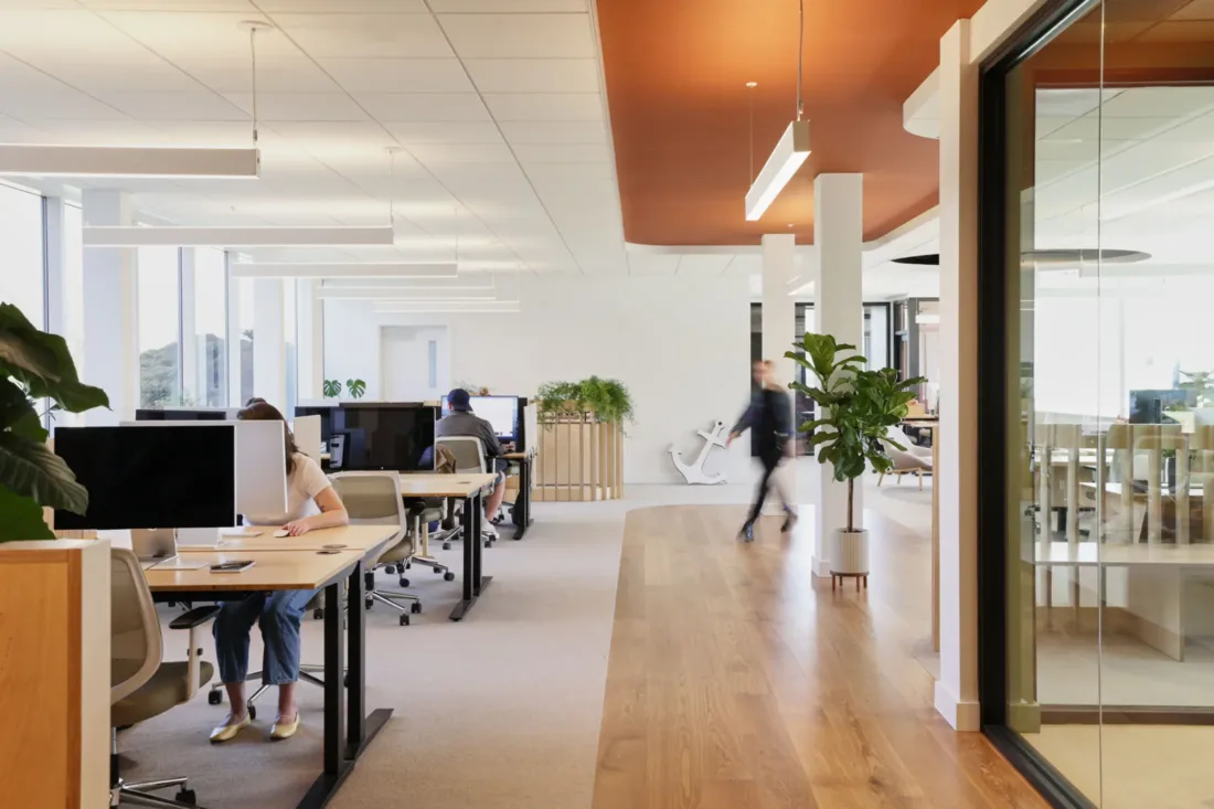 Modern open-plan office with desks, computers, and plants; a person is working at a desk while two people walk through the bright, spacious workspace.