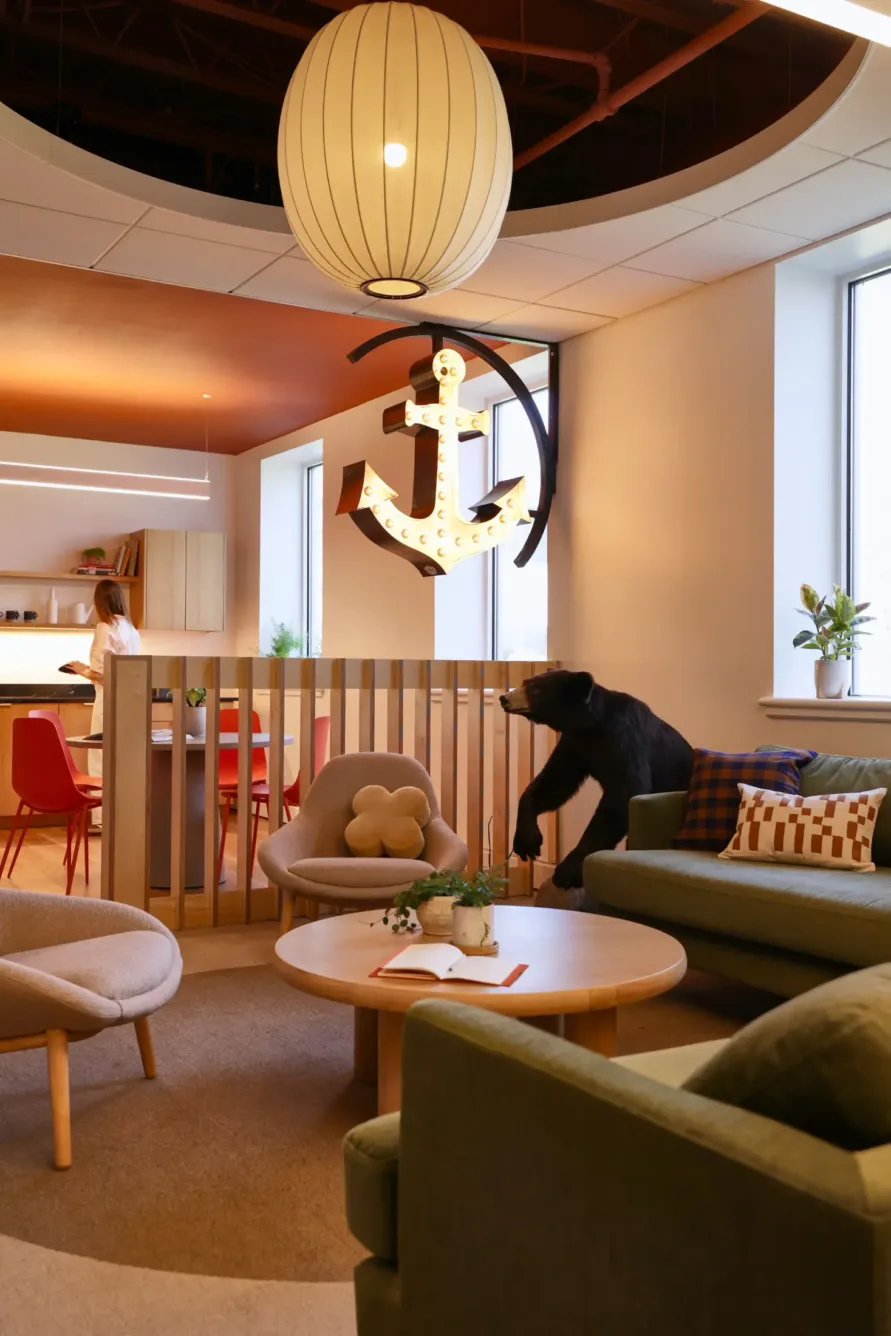 A cozy lounge area with green sofas, a round table, throw pillows, a bear statue, and a large illuminated anchor on the wall. A person stands in the background near kitchen cabinets.