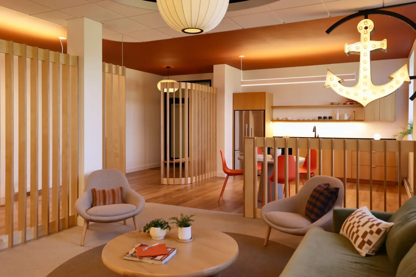 Modern lounge area with cozy chairs, a sofa, round coffee table, potted plants, wooden partitions, open kitchen, and a large illuminated anchor sign hanging from the ceiling.