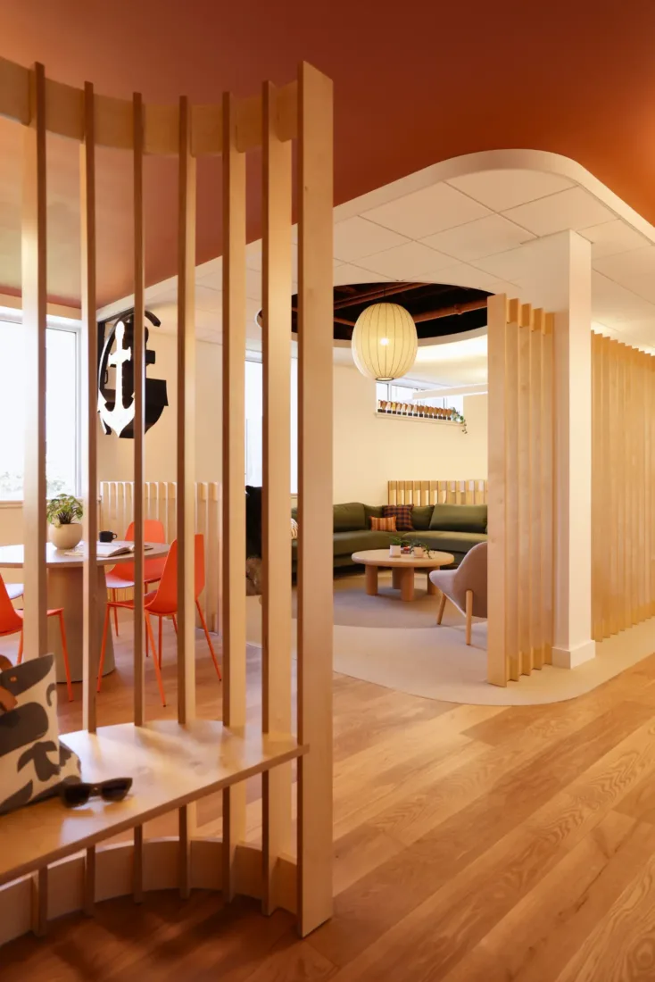 Modern office lounge with wooden slat dividers, orange chairs around round tables, a green sofa, and warm lighting. Large window brings in natural light.