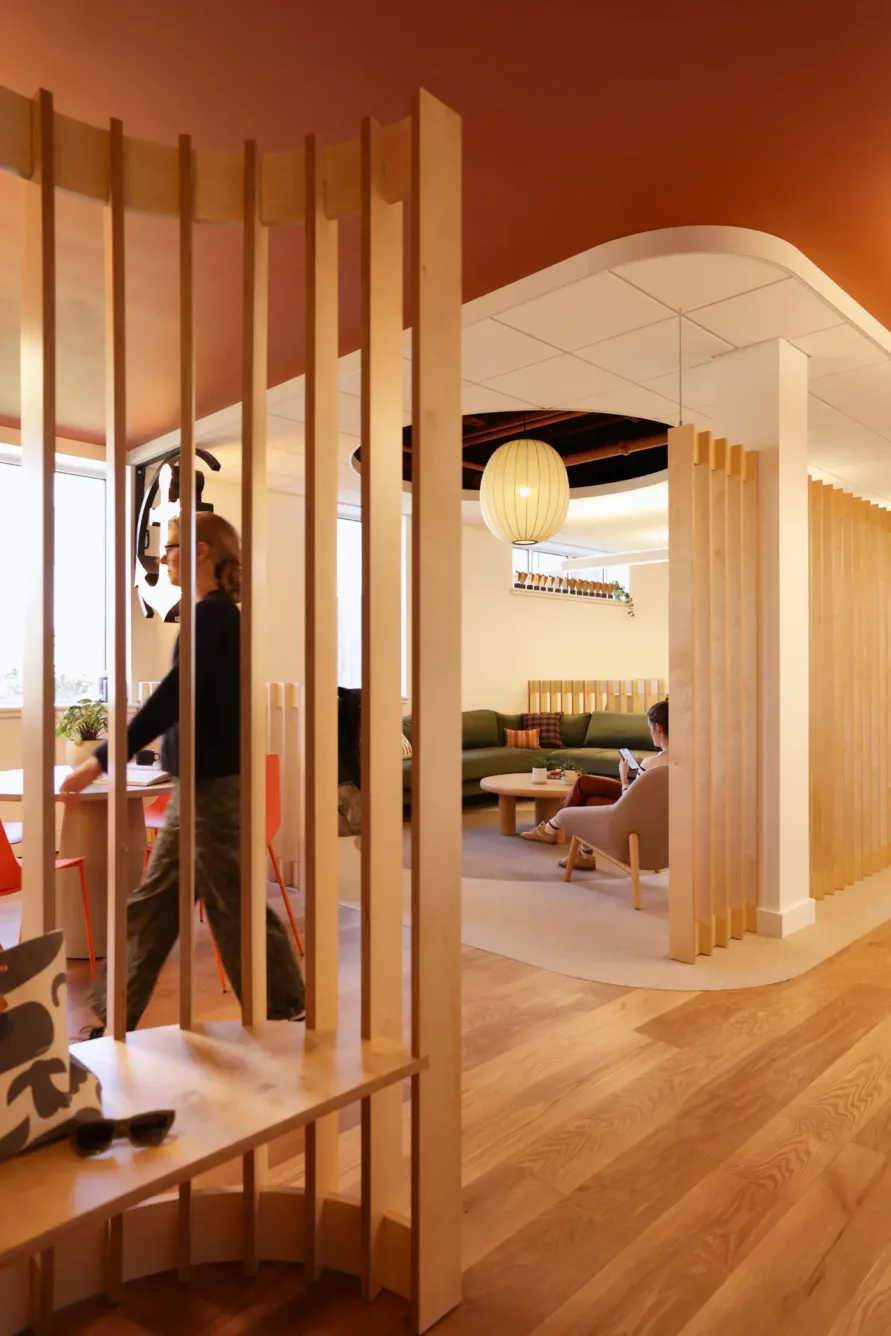 Modern office with wooden partitions, people working and walking, cozy seating, large windows, and warm lighting, creating a welcoming and open workspace atmosphere.