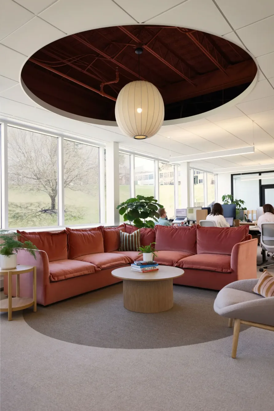 Modern office lounge with pink sectional sofa, round coffee table, plants, large windows, and people working at desks in the background. A round ceiling cutout and pendant light are overhead.