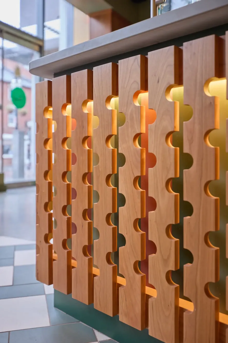 Wooden panel with interlocking puzzle-piece cutouts, backlit with warm yellow lights, beneath a countertop in a modern interior space with windows and tiled floor visible.