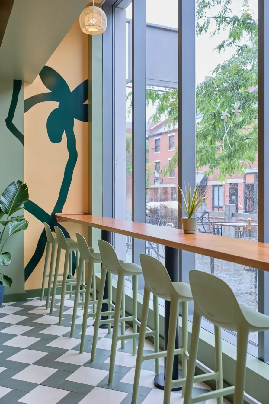 Modern cafe with green chairs, a wood counter by large windows, potted plant, checkered floor, and a mural of an abstract figure on the wall.
