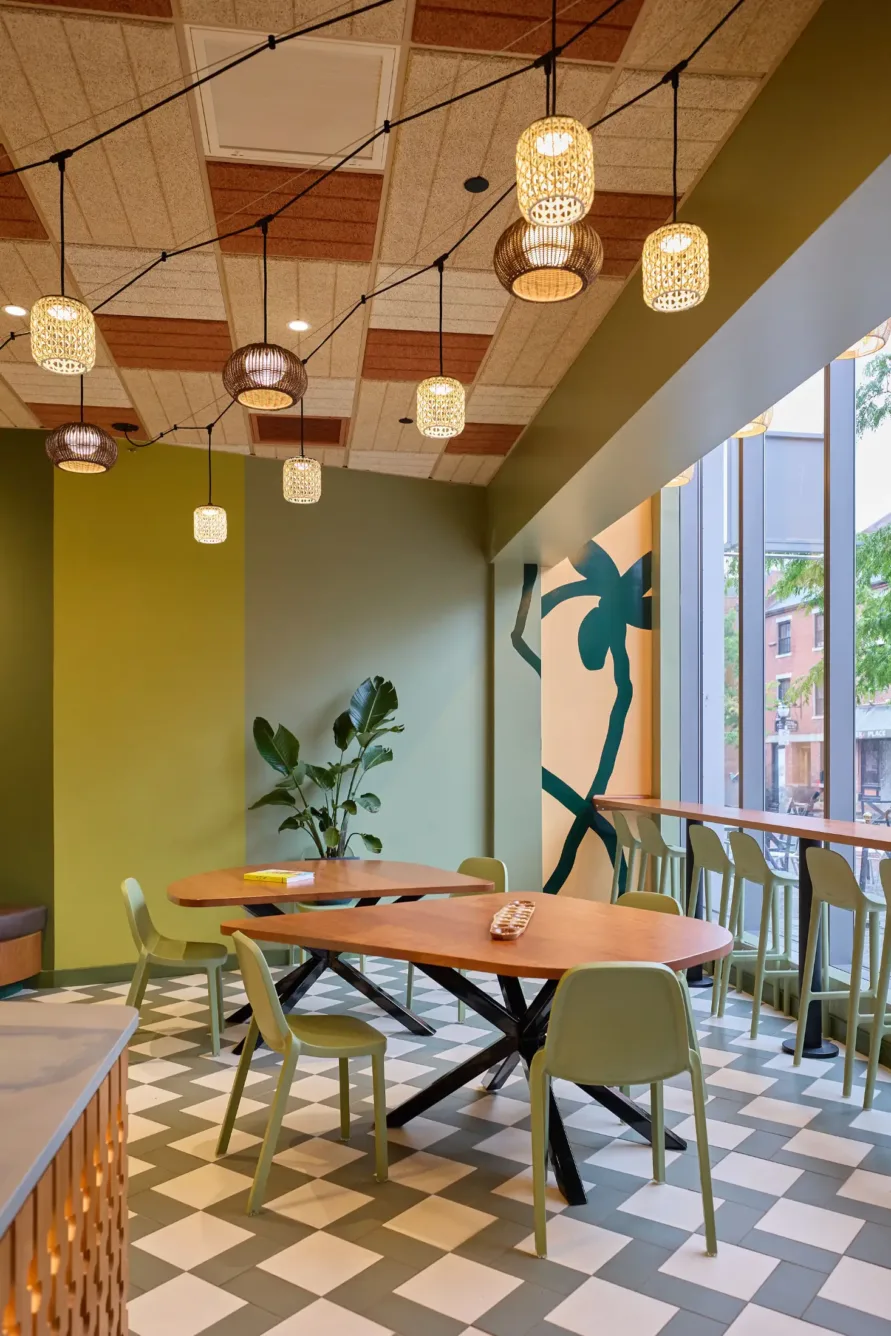 Modern café with green chairs, wooden tables, pendant lights, large windows, and geometric tiled floor. A plant and wall art add a fresh, inviting atmosphere.