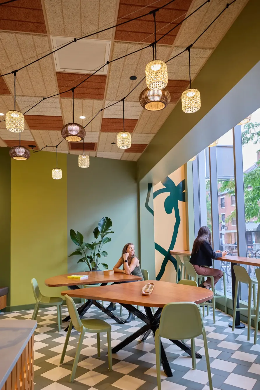 Modern cafe with hanging lights, green walls, large windows, two women working at tables, and a plant mural.