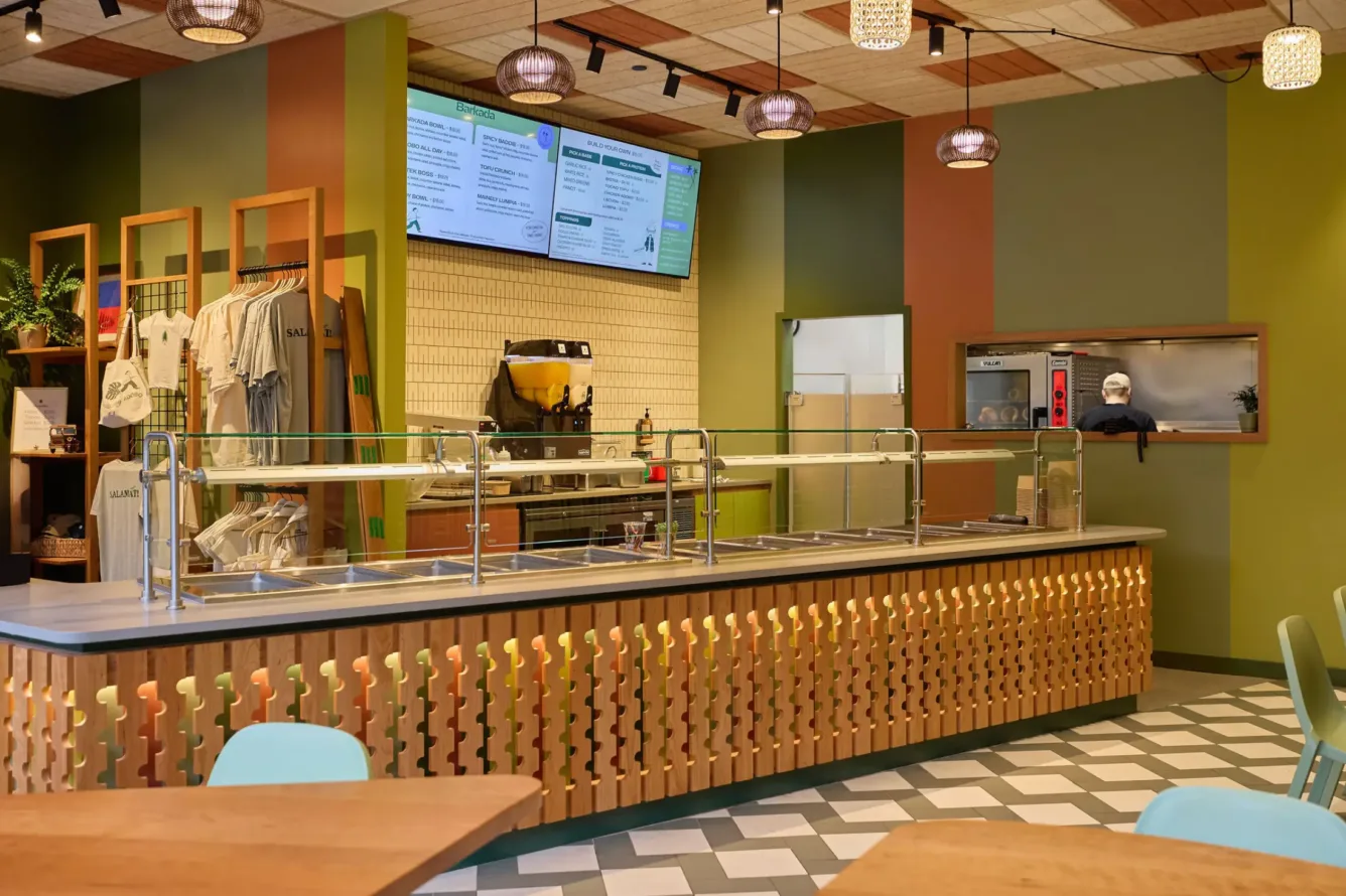 A modern restaurant counter with empty food stations, overhead menus, and a person cooking in the kitchen behind a glass window.