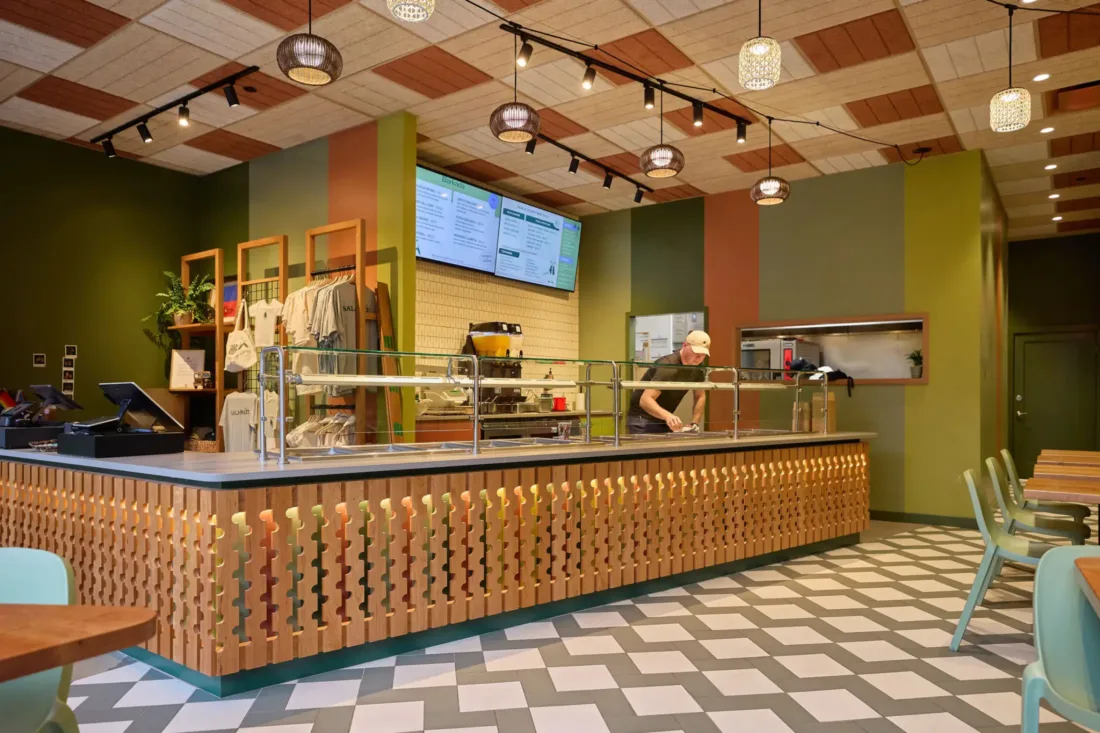 A modern, brightly lit cafe with geometric floor tiles, wooden accents, digital menus, and a staff member preparing food behind the counter.