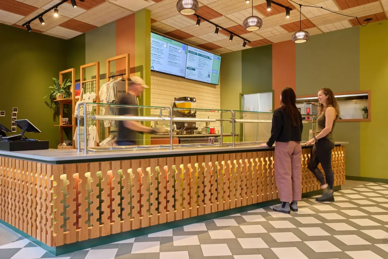 Two women stand at a modern counter with patterned wood and tile flooring, while a worker behind the counter prepares food under menu boards.
