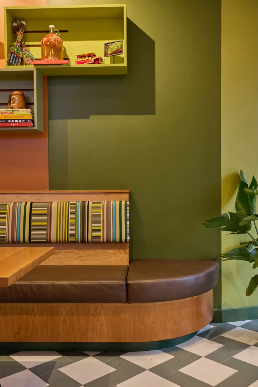 Colorful café booth with striped cushion, wooden table, green walls, geometric floor tiles, shelves with decor, and a potted plant.