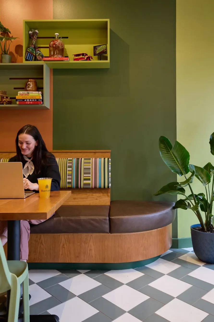 A woman smiles while working on a laptop at a café table with a yellow cup, colorful shelves, and a large potted plant nearby.