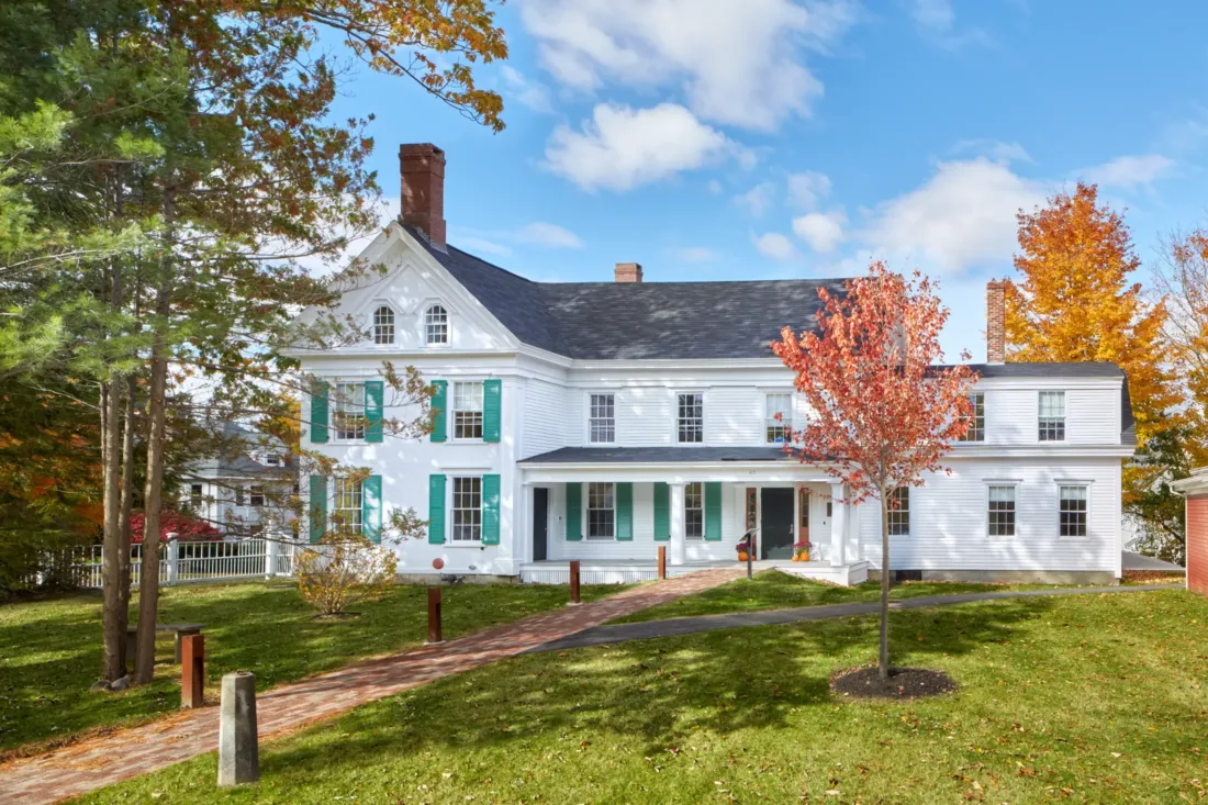 A large white two-story house with green shutters and a front porch, surrounded by grass, trees with autumn leaves, and a brick walkway.