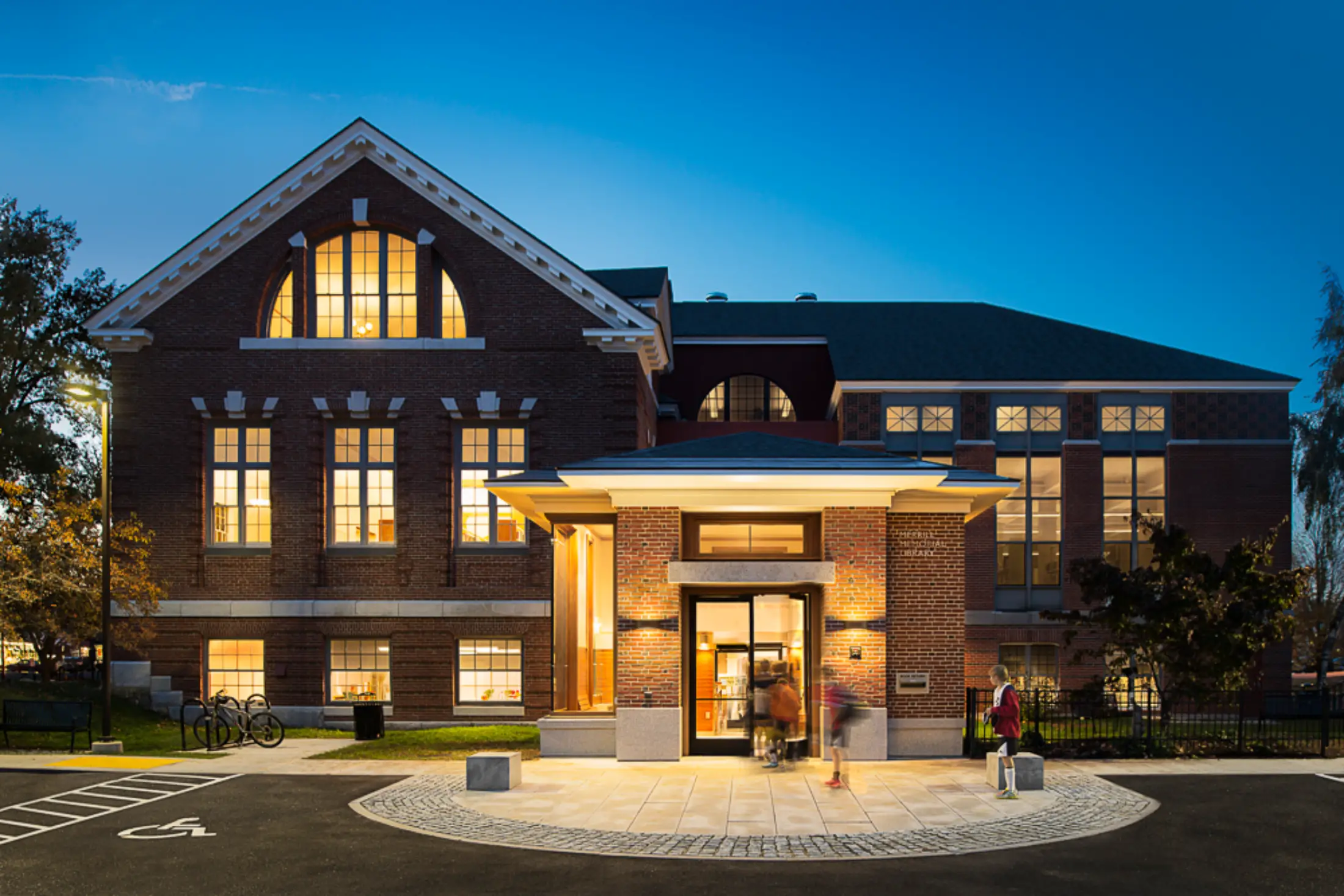 Historic brick building with tall windows lit from inside at dusk; people enter through a brightly lit glass door entrance.