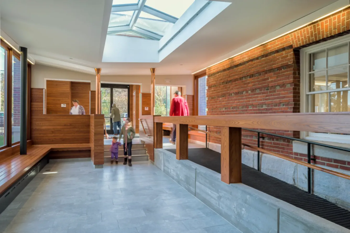 A modern lobby with brick walls, wood benches, skylight, and a wooden ramp; people are walking, including a child holding an adults hand.
