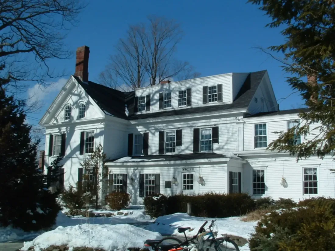 Large white colonial-style house with black shutters, surrounded by snow and trees. Two bicycles are parked in the foreground under clear blue sky.