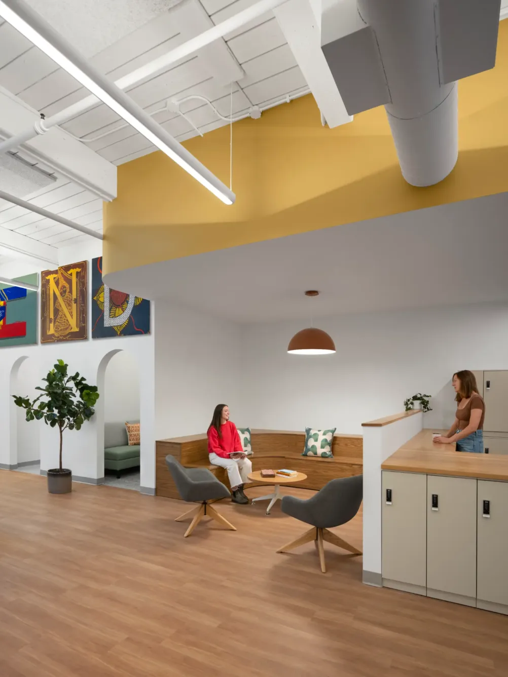 Two women talk in a modern office lounge with wood floors, gray chairs, a plant, artwork on the wall, and yellow ceiling accents.