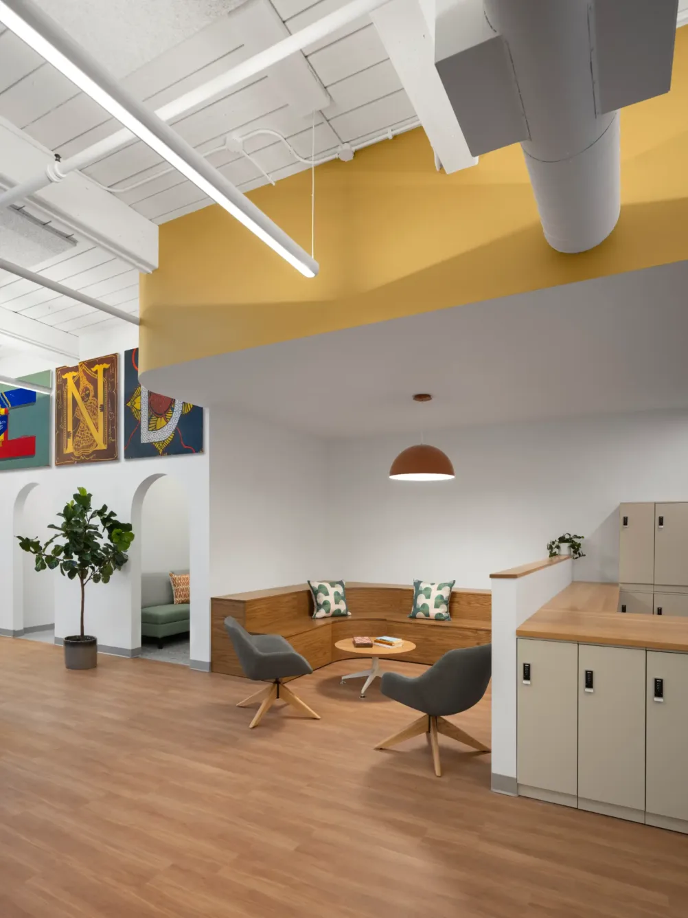 Modern office lounge with wood floors, built-in seating, round table, two gray chairs, lockers, plant, wall art, and overhead lighting.