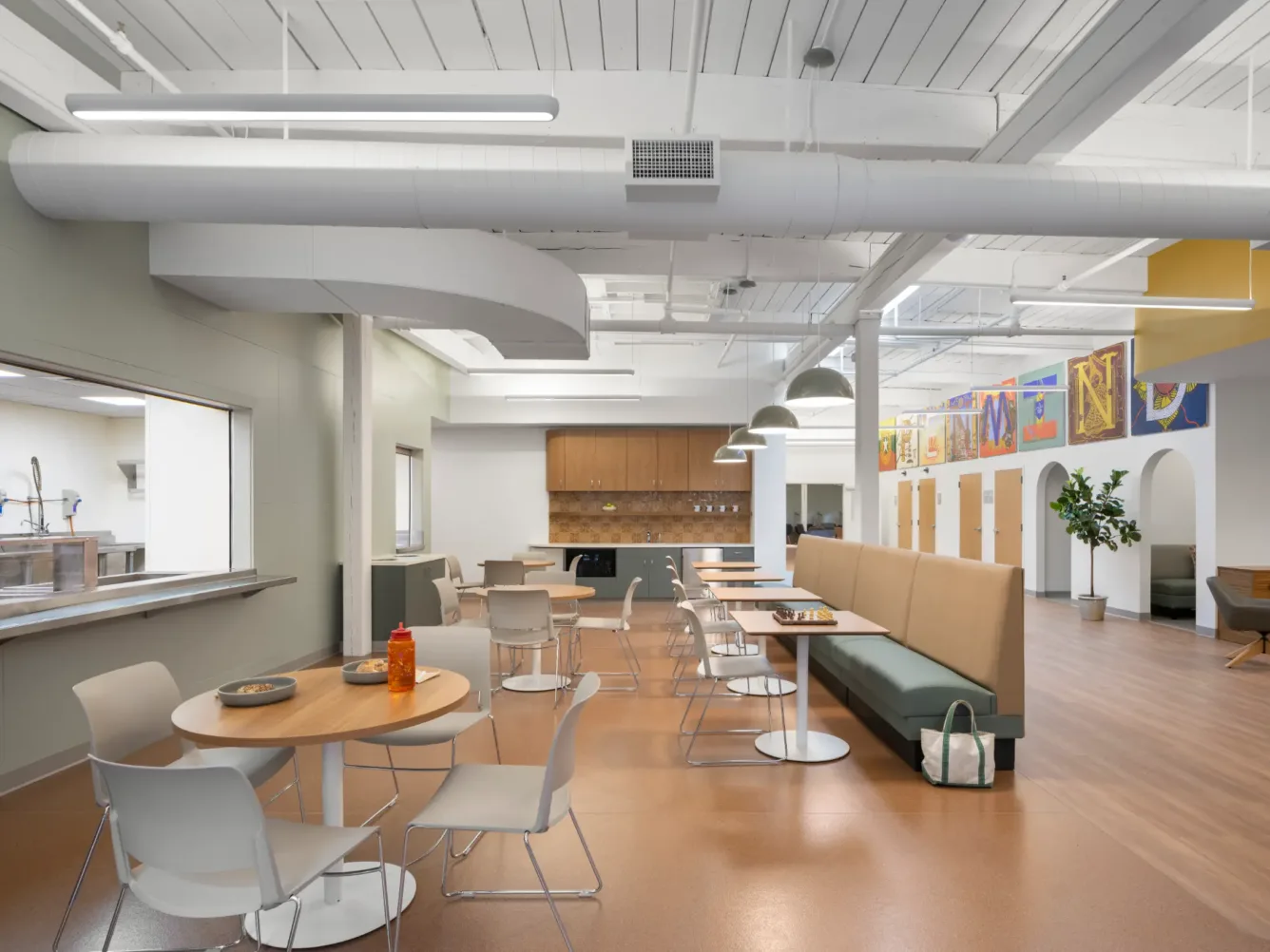Modern cafeteria with round tables, white chairs, a long green bench, and open kitchen. Colorful artwork hangs on the back wall, and large windows let in natural light.