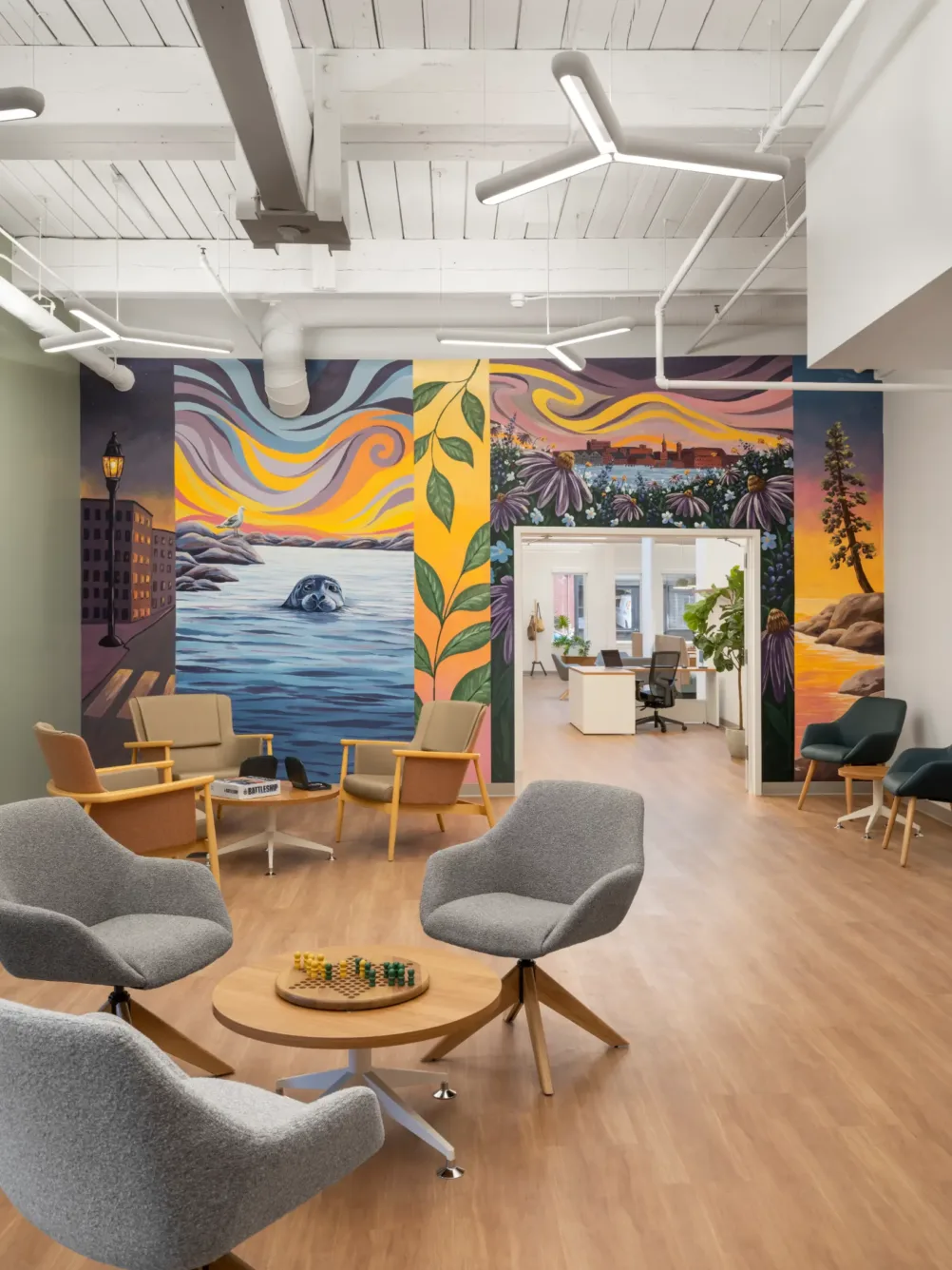 Bright office lounge with gray chairs, a round wood table, and a colorful mural depicting nature and city scenes on the back wall.