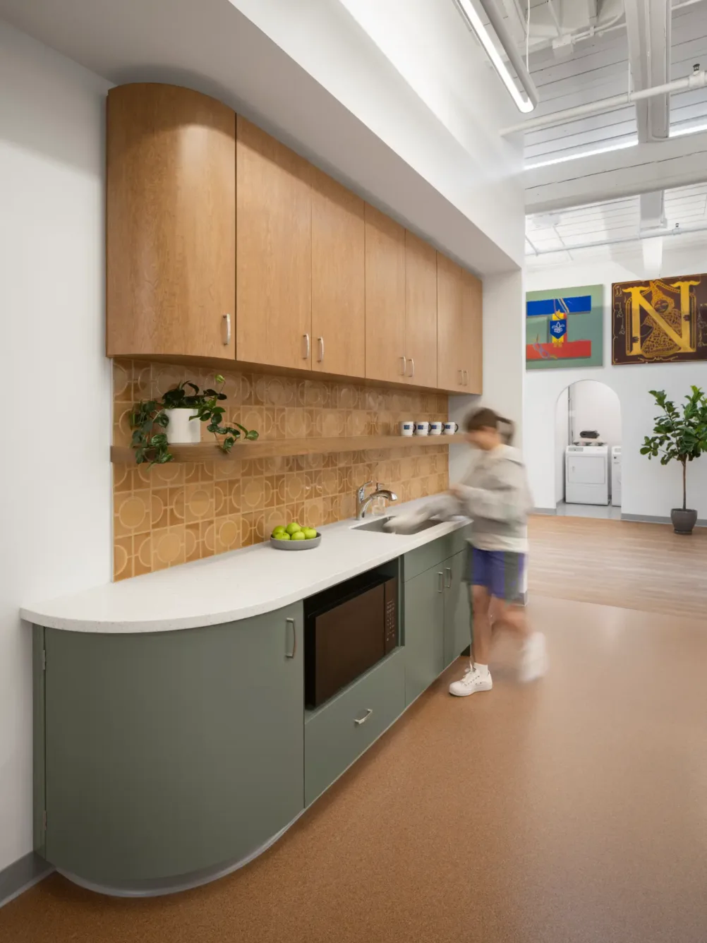 A person stands at a modern kitchen counter with wood cabinets, geometric backsplash, a microwave, and decorative flags on the wall.