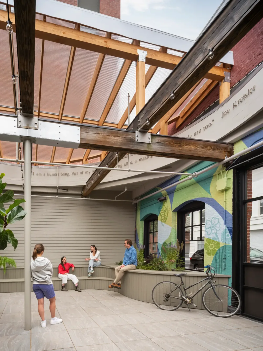Four people sit and talk in a modern outdoor patio with a transparent roof, a mural on the wall, a bicycle, and plants nearby.