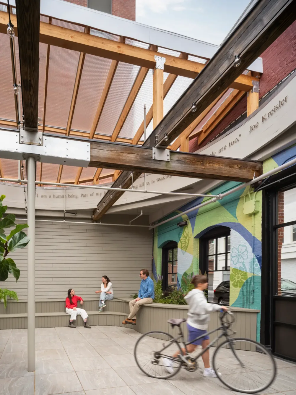 A person rides a bike past three people sitting and talking under a partially covered outdoor patio with a colorful mural on the wall.