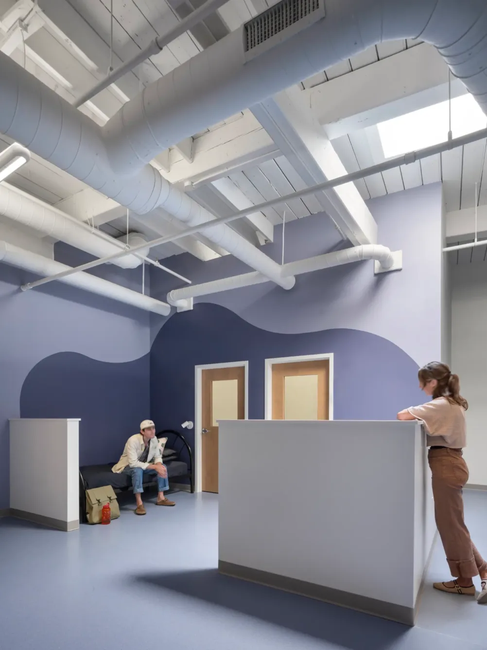 A woman stands at a white reception desk while a man waits on a bench. The room has exposed pipes, skylights, and blue-purple wavy wall décor.