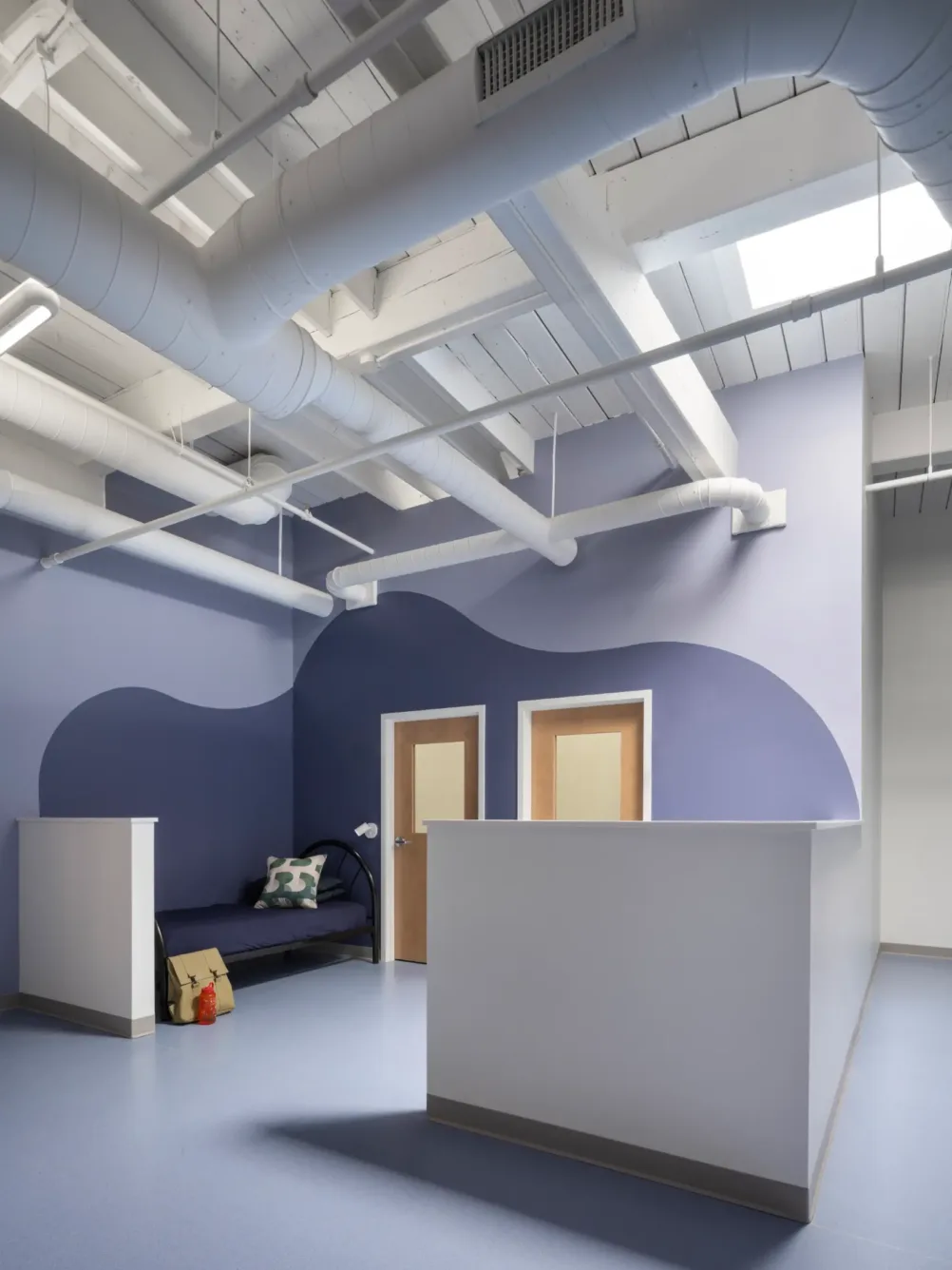 Modern room with exposed white pipes, blue wavy wall design, two wooden doors, a dark sofa with pillows, and a red bag on the floor. Skylight brings in natural light.