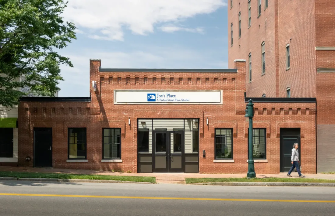 A person walks past a brick building with a sign over the door reading Joes Place Public Tavern Since Twelve. The sky is partly cloudy and a tree is on the left side.