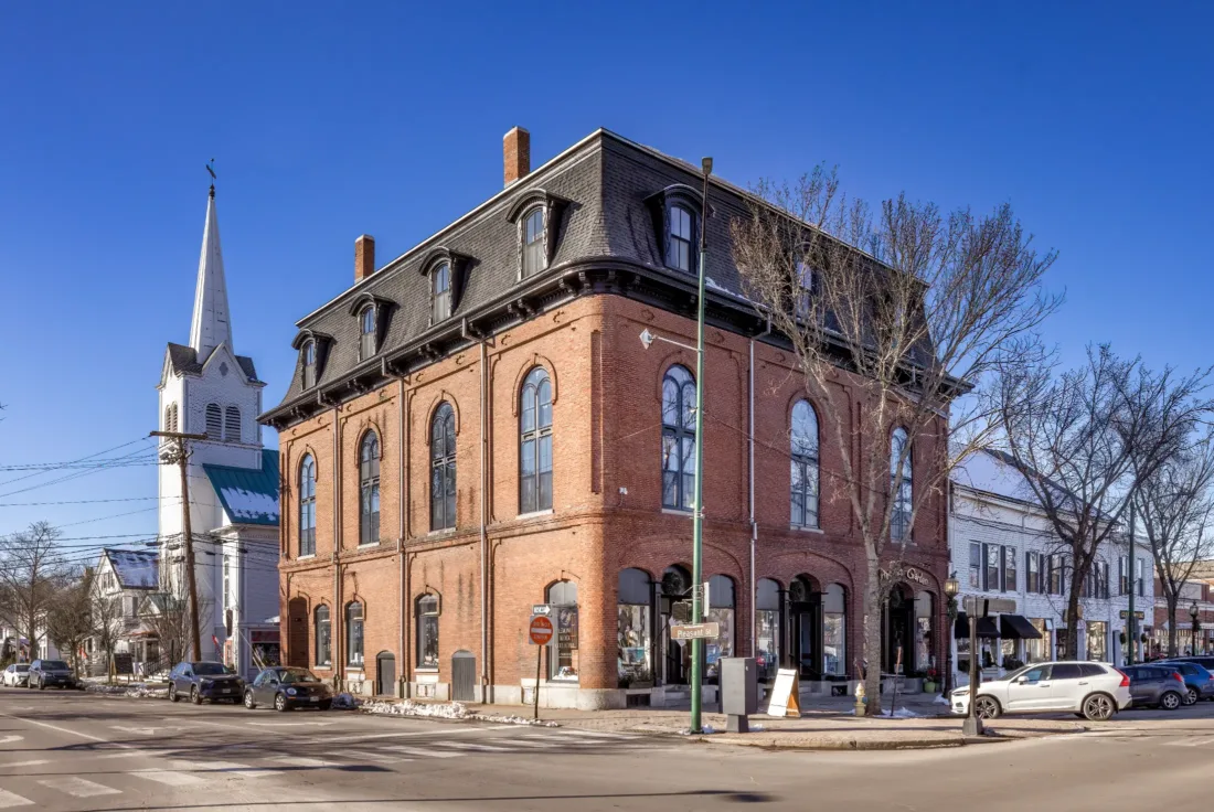 Historic red brick building with arched windows on a street corner; church with steeple and shops visible in the background, under a clear blue sky.