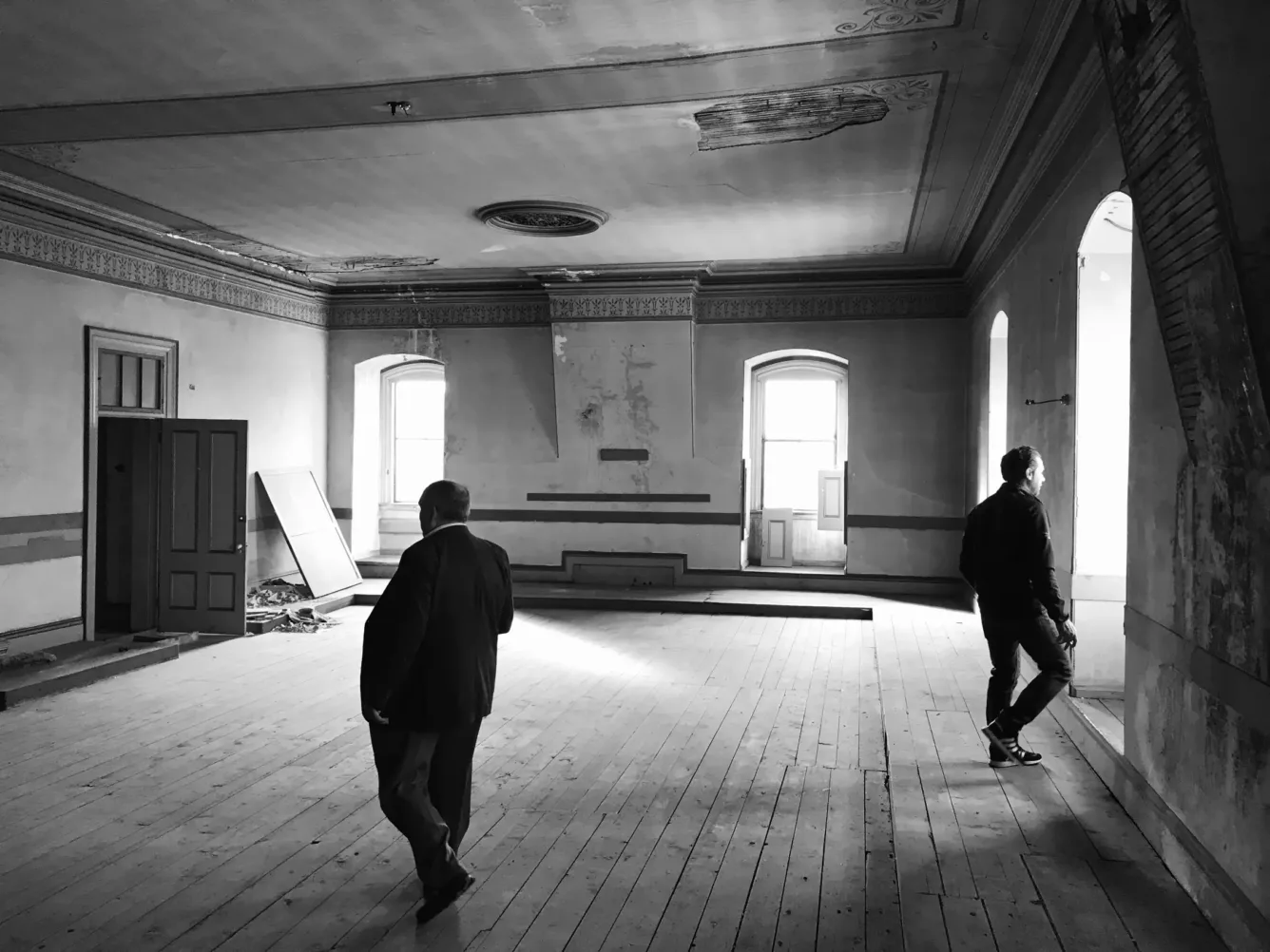 Two men stand in a large, empty, old room with wooden floors and damaged ceiling. Light comes in from tall windows.